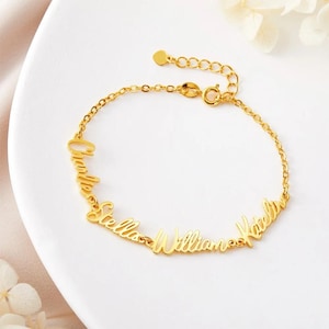Puede incluir: Una pulsera dorada con los nombres "Charlotte", "Stella", "William" y "Katelyn" en escritura cursiva. La pulsera tiene una cadena ajustable y se muestra sobre una superficie blanca.