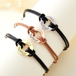 Op de afbeelding: Drie gepersonaliseerde armbanden met ronde hangers. Elke hanger is gegraveerd met een naam en een pootafdrukontwerp met vleugels. De armbanden hebben zwarte en bruine leren koorden. Eén hanger is goudkleurig, één is roségoud en één is zilver.