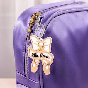 Può includere: Un portachiavi a tema balletto con un fiocco bianco, scarpette da ballo e il nome "Ella Renee" su uno striscione. Il portachiavi è attaccato a uno zaino viola. Un piccolo ciondolo a forma di ballerina dorata pende dal fermaglio.