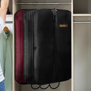 Puede incluir: Tres fundas para prendas colgadas en un armario. Las fundas son negras, rojas y negras con rayas verticales. La bolsa negra tiene el nombre "BARNETT" impreso en dorado. Las fundas cuelgan de una barra met&aacute;lica.