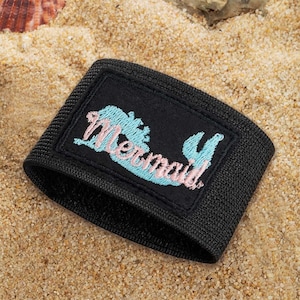 May include: A black elastic armband featuring a rectangular patch with the word Mermaid embroidered in light blue and pink thread. The text is stylised with a cursive font and sits against a solid black background on the fabric band.
