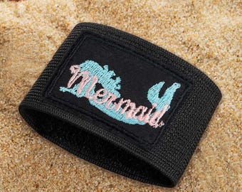 May include: A black elastic armband featuring a rectangular patch with the word Mermaid embroidered in light blue and pink thread. The text is stylized with a cursive font and sits against a solid black background on the fabric band.