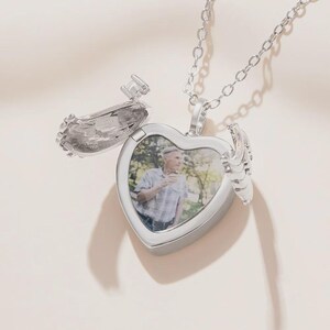 May include: A silver heart-shaped locket necklace with an openable compartment. The locket features a photo inside and wing-like details on the sides. The chain is also silver and hangs from the top of the heart.