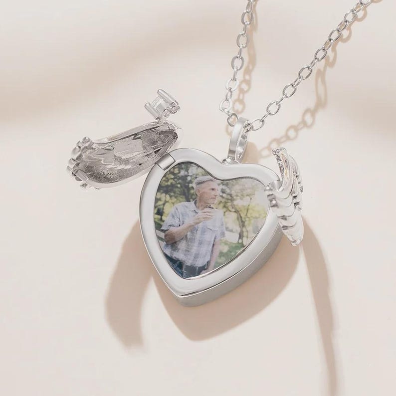 May include: A silver heart-shaped locket necklace with an openable compartment. The locket features a photo inside and wing-like details on the sides. The chain is also silver and hangs from the top of the heart.