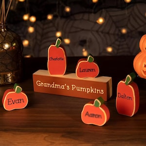 Könnte beinhalten: Ein Holzschild mit dem Text "Grandma's Pumpkins" und fünf kleine, rote, hölzerne Kürbisse mit Namen darauf: Charlotte, Lauren, Evan, Dalton und Aaron.