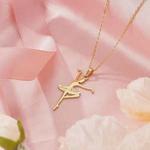 Könnte beinhalten: Eine goldfarbene Halskette mit einem Ballerina-Anhänger. Der Anhänger zeigt eine Ballerina in einer anmutigen Pose, mit dem eingravierten Namen "Eva-Rose". Die Halskette liegt vor einem rosa Satinhintergrund.