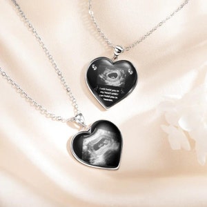 May include: Two silver necklaces with heart-shaped pendants. Each pendant features a black and white ultrasound image of a baby. The pendant on the top necklace has the text "I will hold you in my heart until I can hold you in heaven."