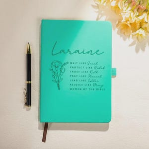 Könnte beinhalten: Ein türkisfarbenes Notizbuch mit dem Namen "Laraine" in Schreibschrift. Das Notizbuch zeigt florale Linienkunst und den Text: "Warte wie Sarah, Beschütze wie Rahab, Vertraue wie Ruth, Bete wie Hannah, Führe wie Esther, Freue dich wie Maria, Frauen der Bibel." Ein schwarzer Stift mit goldenen Akzenten liegt daneben.