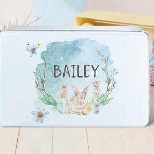 May include: A white metal tin with a blue watercolor design featuring a wreath of flowers and three bunnies. The name "BAILEY" is written in black lettering on the tin.