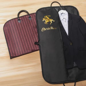 Puede incluir: Funda de traje negra con un jinete bordado en oro y el nombre "Christian". Un traje negro y una camisa blanca cuelgan dentro. Tambi&eacute;n se ve una funda de ropa a rayas burdeos y negras con un asa.