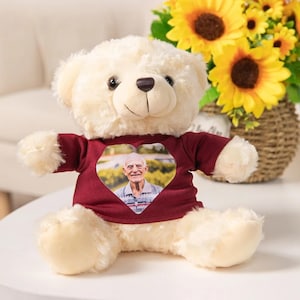 Puede incluir: Un oso de peluche blanco que lleva una camisa granate con una foto en forma de corazón de un hombre mayor. El oso está sentado sobre una superficie blanca.