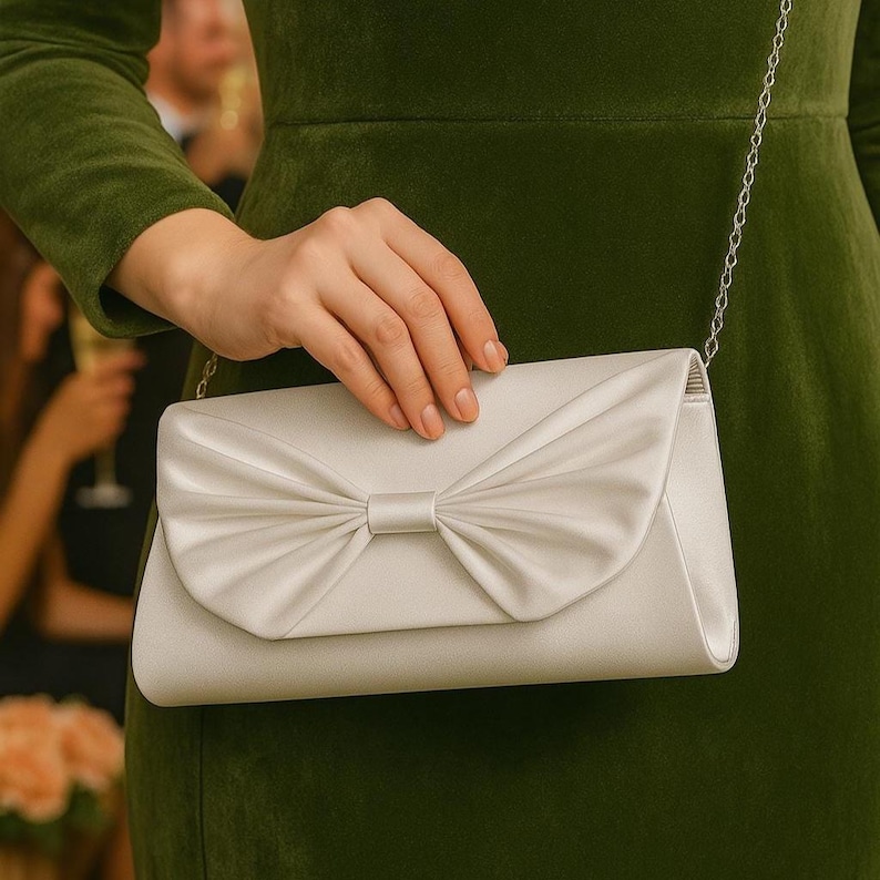 K&ouml;nnte beinhalten: Eine satin Clutch in hellem Elfenbein, mit einer gro&szlig;en Schleife auf der Vorderseite. Die Clutch hat einen silbernen Kettenriemen und eine rechteckige Form. Im Hintergrund ist eine Person in einem gr&uuml;nen Kleid zu sehen.