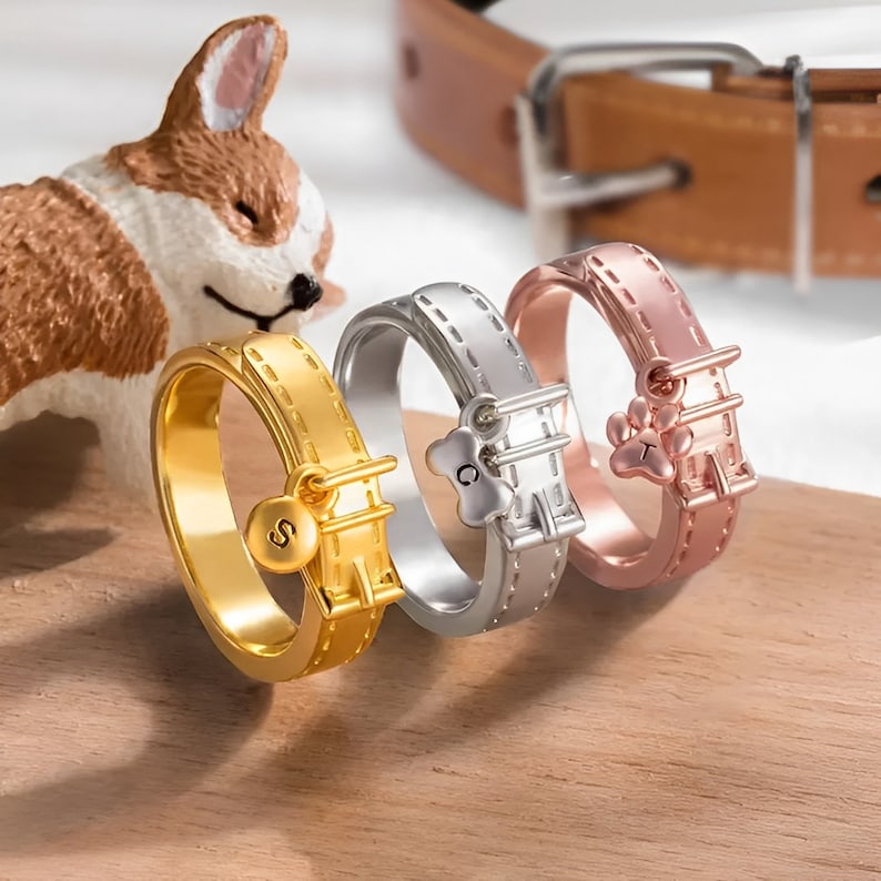 May include: Three gold, silver, and rose gold rings shaped like dog collars with charms. The gold ring has a charm with the letter "S", the silver ring has a charm with the letter "C" and a paw print, and the rose gold ring has a charm with the letter "T" and a paw print.