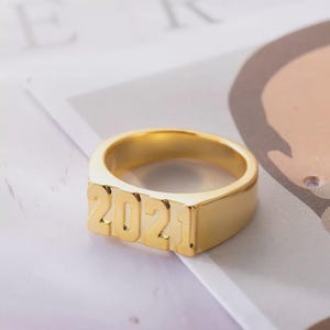 以下が含まれることがあります： 数字「2021」が浮き彫りになったゴールドカラーのリング。リングは長方形の顔と磨かれたバンドを持っています。リングはニュートラルな背景に配置されています。