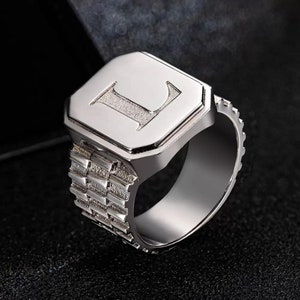 Puede incluir: Un anillo de plata con una parte superior de forma cuadrada que presenta una letra "L" en relieve. La banda del anillo tiene un diseño texturizado y estampado.