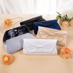 K&ouml;nnte beinhalten: Eine Auswahl an Satin-Clutch-Taschen in verschiedenen Farben, darunter Schwarz, Marineblau und Creme. Eine Tasche ist ge&ouml;ffnet und zeigt ein Schwarz-Wei&szlig;-Foto. Jede Clutch hat eine gro&szlig;e Schleife auf der Vorderseite, passend f&uuml;r Hochzeiten oder besondere Anl&auml;sse.