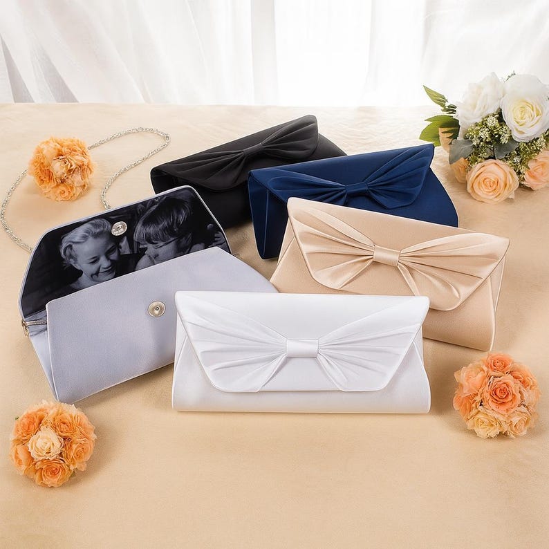 K&ouml;nnte beinhalten: Eine Auswahl an Satin-Clutch-Taschen in verschiedenen Farben, darunter Schwarz, Marineblau und Creme. Eine Tasche ist ge&ouml;ffnet und zeigt ein Schwarz-Wei&szlig;-Foto. Jede Clutch hat eine gro&szlig;e Schleife auf der Vorderseite, passend f&uuml;r Hochzeiten oder besondere Anl&auml;sse.