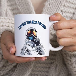 Op de afbeelding: Een witte keramische mok met een afbeelding van een skiër die een helm, skibril en gezichtsmasker draagt, met een vinger op de lippen in een stiltegebaar. De tekst boven de illustratie luidt, I SKI LIKE YOU WISH YOU COULD.