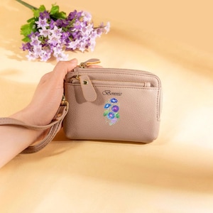 May include: A taupe-colored wallet with a zipper closure and a wrist strap. The wallet features the embroidered word "Bonnie" above a floral design with blue, purple, and green accents. A sprig of purple flowers is in the background.