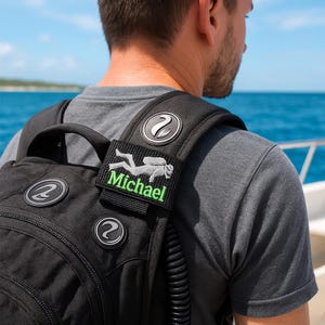 Op de afbeelding: Zwarte rugzak met een duikthema. Voorzien van een patch met een duiker silhouet en de naam "Michael" in het groen. Inclusief zilverkleurige ronde emblemen met een gestileerd "2" ontwerp.
