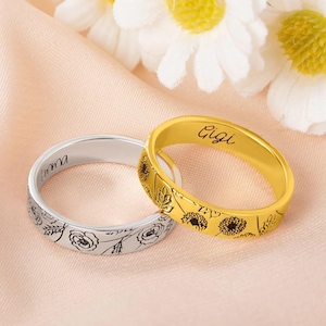 Puede incluir: Dos anillos, uno plateado y otro dorado, sobre una superficie rosa. El anillo plateado tiene un diseño floral y el nombre "Emma". El anillo dorado tiene el nombre "Gigi" y un patrón floral.