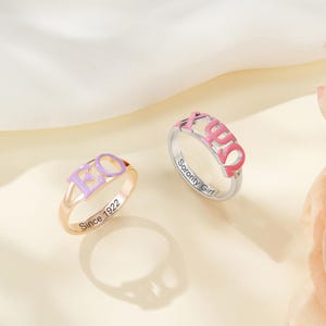 May include: Two rings with sorority letters. One ring is gold-toned with lavender letters and the text "Since 1922". The other ring is silver-toned with pink letters and the text "Sorority Girl".