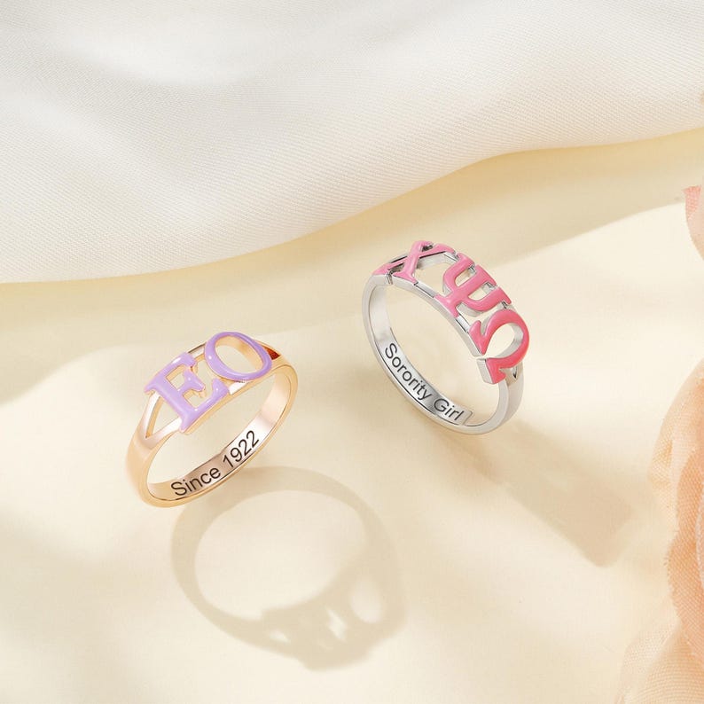 May include: Two rings with sorority letters. One ring is gold-toned with lavender letters and the text "Since 1922". The other ring is silver-toned with pink letters and the text "Sorority Girl".