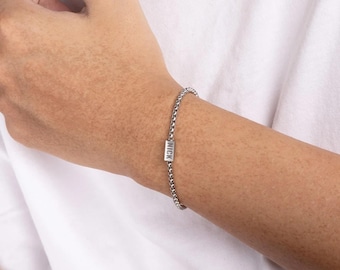 May include: A silver-colored bracelet with a chain-link design. The bracelet features a rectangular charm with the name "NICK" engraved on it. The bracelet is worn on a wrist against a white shirt.