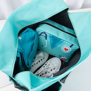 May include: A teal blue travel bag with a black zipper and a black mesh pocket. The bag is open and contains a teal blue pouch with a stethoscope and heart graphic, a teal blue shirt, and two white sneakers.