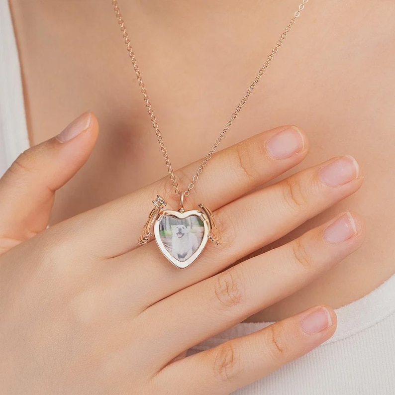 May include: A rose gold heart-shaped locket necklace with a photo of a dog inside. The locket has small wings on either side and hangs from a delicate chain. The necklace is being held by a person.