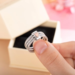 Pu&ograve; includere: Un anello d'argento a tre fasce, con un piccolo accento a forma di cuore al centro. L'anello &egrave; inciso con i nomi "Sophia", "Michael" e "Levi". L'anello &egrave; tenuto davanti a uno sfondo neutro.