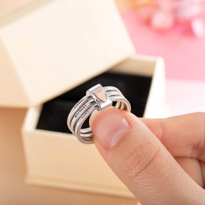 Pu&ograve; includere: Un anello d'argento a tre fasce, con un piccolo accento a forma di cuore al centro. L'anello &egrave; inciso con i nomi "Sophia", "Michael" e "Levi". L'anello &egrave; tenuto davanti a uno sfondo neutro.