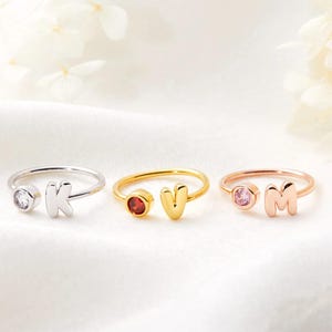May include: Three open-style rings with initial letters and gemstones. The rings are silver, gold, and rose gold. The silver ring has a clear gemstone and the letter "K". The gold ring has a red gemstone and the letter "V". The rose gold ring has a pink gemstone and the letter "M".