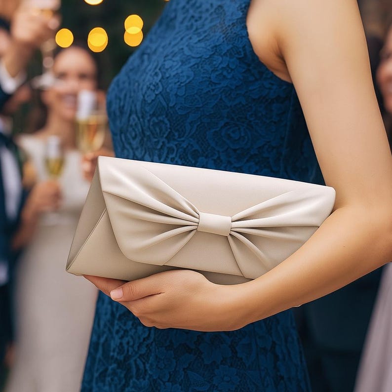 K&ouml;nnte beinhalten: Eine cremefarbene Clutch mit Schleifendetail. Die rechteckige Tasche wird von einer Person getragen, die ein blaues Spitzenkleid tr&auml;gt. Der Hintergrund zeigt eine verschwommene Outdoor-Veranstaltung mit Menschen und Lichtern.
