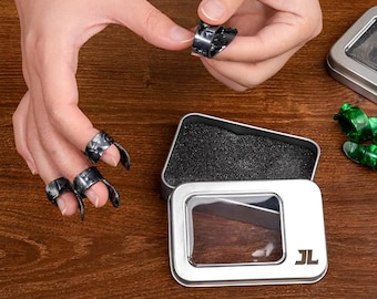 May include: Set of silver-colored finger picks for guitar or other stringed instruments. The picks are in a small metal tin with a clear window and the initials "JL". Some picks are on the fingers of a person.