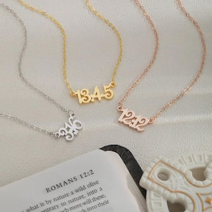 May include: Four delicate necklaces with numbers, in silver, gold, and rose gold. The numbers are 3:16, 13.4.5, and 12.12. The necklaces are on a light surface, with a book open to Romans 12:2 in the background.
