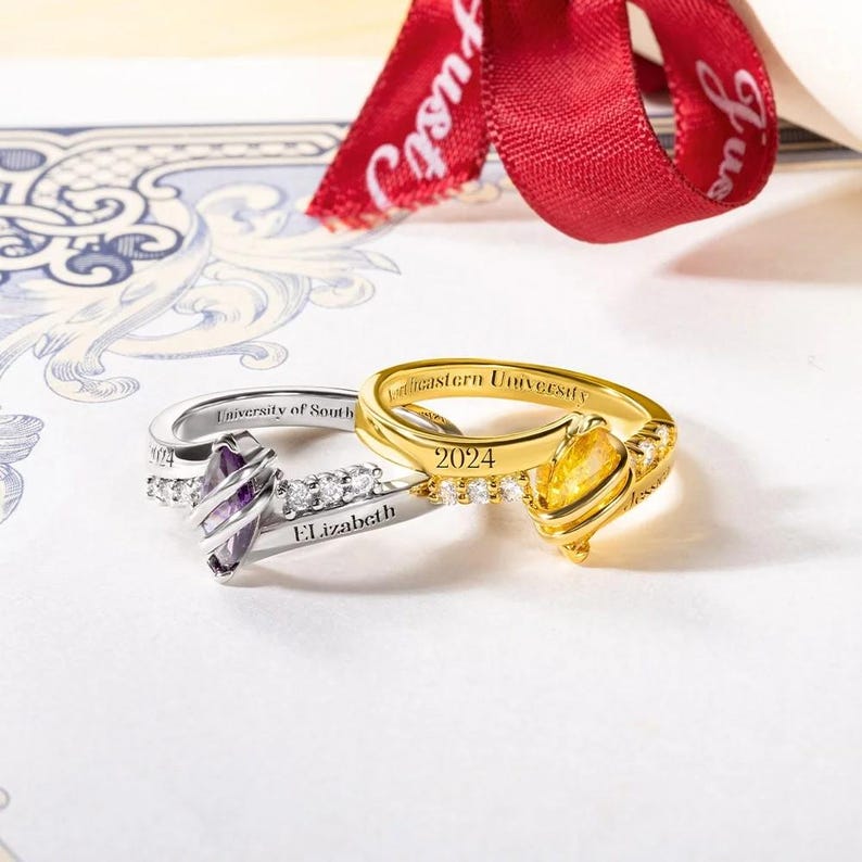 May include: Two gold and silver rings with engraved text. The silver ring has a purple stone and the text "University of South Florida Elizabeth". The gold ring has a yellow stone and the text "Northeastern University 2024".
