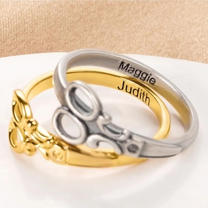 Puede incluir: Dos anillos, uno dorado y otro plateado, apilados. El anillo dorado tiene un diseño decorativo en la parte delantera. El anillo plateado tiene grabados los nombres "Maggie" y "Judith". Los anillos están sobre una superficie blanca.