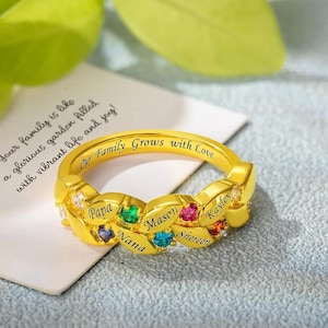 May include: Gold ring with a braided design and colorful gemstones. The ring is engraved with the words "Our Family Grows with Love" and the names "Papa", "Nana", "Masen", "Shereen", and "Kayley".