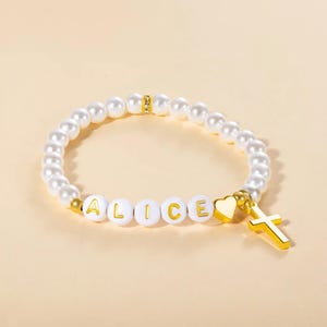Könnte beinhalten: Ein weißes Perlenarmband mit dem Namen "ALICE" in weißen Buchstabenperlen. Das Armband hat einen goldenen Herzanhänger und einen goldenen Kreuzanhänger. Das Armband besteht aus Kunstperlen.