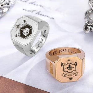 Puede incluir: Dos anillos de sello. Uno es plateado con un diseño de escudo de armas en blanco y negro y el nombre "ERICK" grabado en el lateral. El otro es dorado con un diseño de escudo de armas negro y el año "2007" grabado en el lateral. Ambos anillos tienen texto.