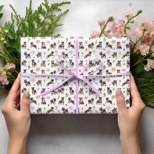 May include: A white gift box with a brown and white dog pattern is wrapped with a light purple ribbon. The box is surrounded by green leaves and pink flowers.