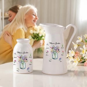 Puede incluir: Juego de jarra y jarrón de cerámica blanca. Cada pieza presenta un diseño floral en un tarro de cristal con el texto "Mum's Garden". La jarra tiene un asa y un pico. El jarrón es de forma pequeña y redondeada.