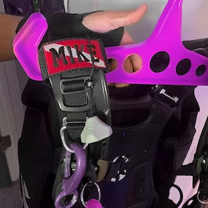 Op de afbeelding: Een zwarte duik-BCD (Buoyancy Control Device) met een rood-wit embleem met de tekst "MIKE". De BCD heeft een paarse karabijnhaak en een wit-grijs label. Een felroze kleerhanger is ook zichtbaar.