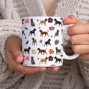 May include: White ceramic mug with a repeating pattern of dogs, flowers, and leaves in brown, black, and pink. The mug has a white handle. The mug is held by a person wearing a cream-coloured sweater.