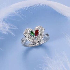 Puede incluir: Un anillo plateado con un dise&ntilde;o en forma de coraz&oacute;n, con una piedra preciosa roja y verde, y peque&ntilde;as piedras blancas. El anillo tiene un motivo floral y la palabra "Always" est&aacute; grabada en la banda. El fondo es azul claro.