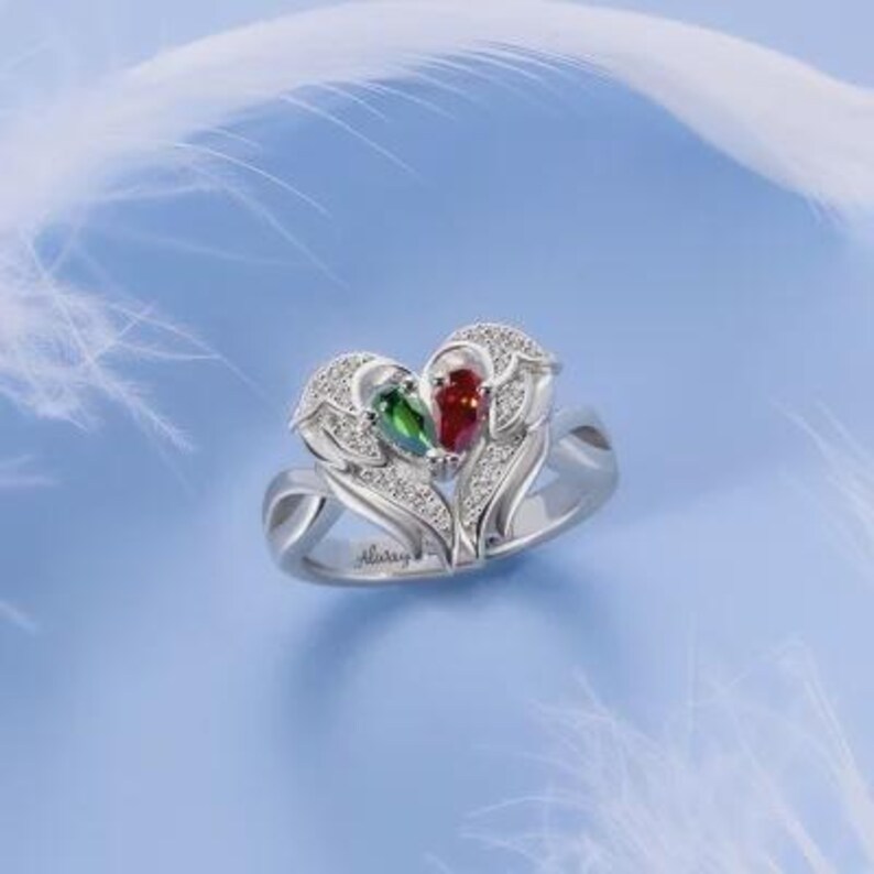 Puede incluir: Un anillo plateado con un dise&ntilde;o en forma de coraz&oacute;n, con una piedra preciosa roja y verde, y peque&ntilde;as piedras blancas. El anillo tiene un motivo floral y la palabra "Always" est&aacute; grabada en la banda. El fondo es azul claro.