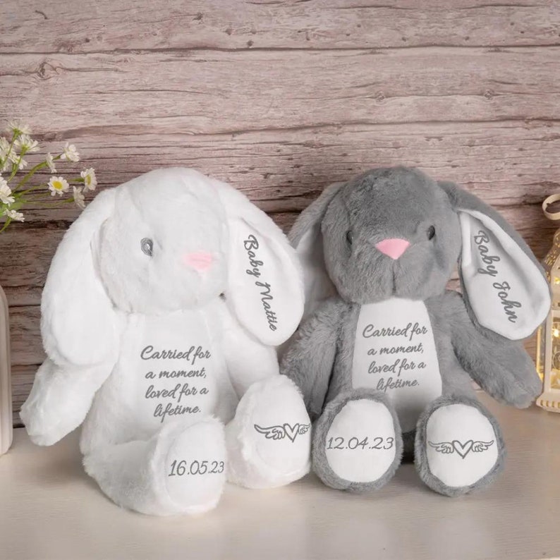 May include: Two plush bunny rabbits, one white and one gray, with embroidered details. The white bunny has "Baby Mattie" on its ear and "16.05.23" on its foot. The gray bunny has "Baby John" on its ear and "12.04.23" on its foot. Both bunnies have the text "Carried for a moment, loved for a lifetime."
