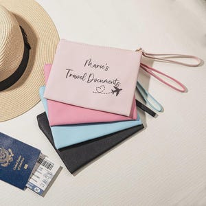 May include: A stack of four travel document pouches in pink, light blue, and black, with a straw hat in the background. The top pouch is pink and has the text "Marie's Travel Documents" with a heart and airplane graphic.