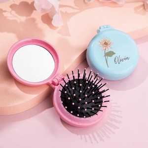 Peut inclure: Un miroir compact rose avec une petite brosse au dos. Le miroir a un fond blanc et la brosse a des poils noirs. Le compact est ouvert et le miroir est visible. Le compact est sur une surface rose. Le compact porte le nom "Olivia".